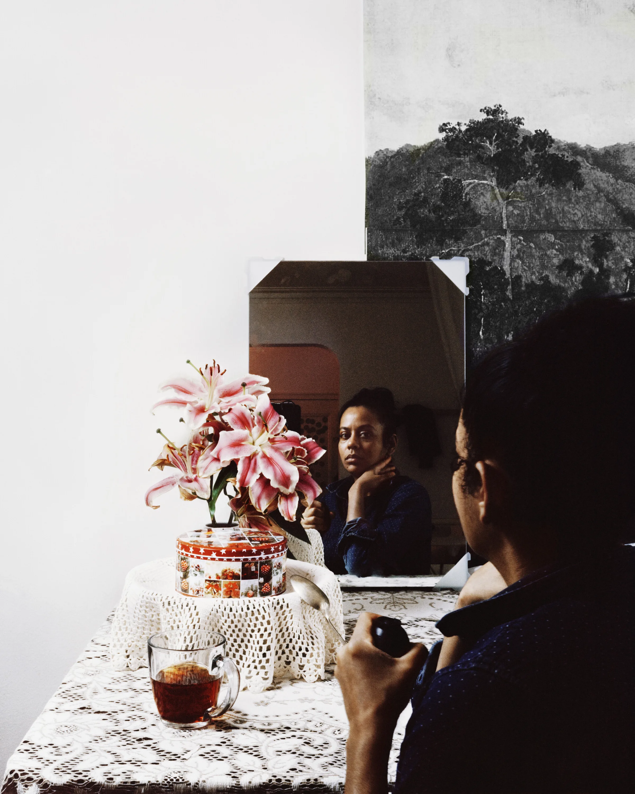 A dark-skinned woman sits looking at her reflection in the mirror with one hand supporting her chin. In front of her are pink flowers, a red and white round tin box, and a clear mug with dark brown liquid.