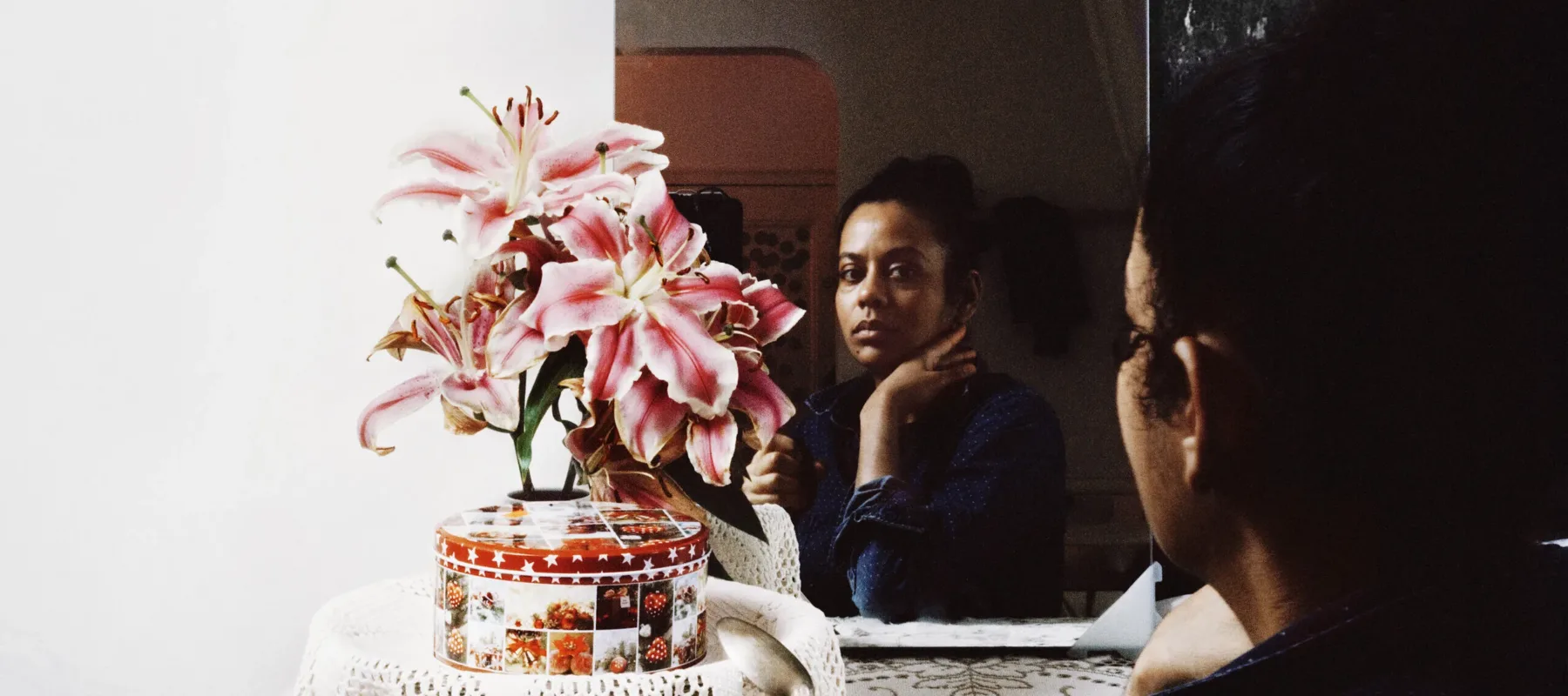 A dark-skinned woman sits looking at her reflection in the mirror with one hand supporting her chin. In front of her are pink flowers, a red and white round tin box, and a clear mug with dark brown liquid.