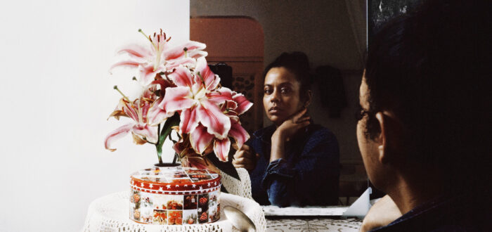 A dark-skinned woman sits looking at her reflection in the mirror with one hand supporting her chin. In front of her are pink flowers, a red and white round tin box, and a clear mug with dark brown liquid.