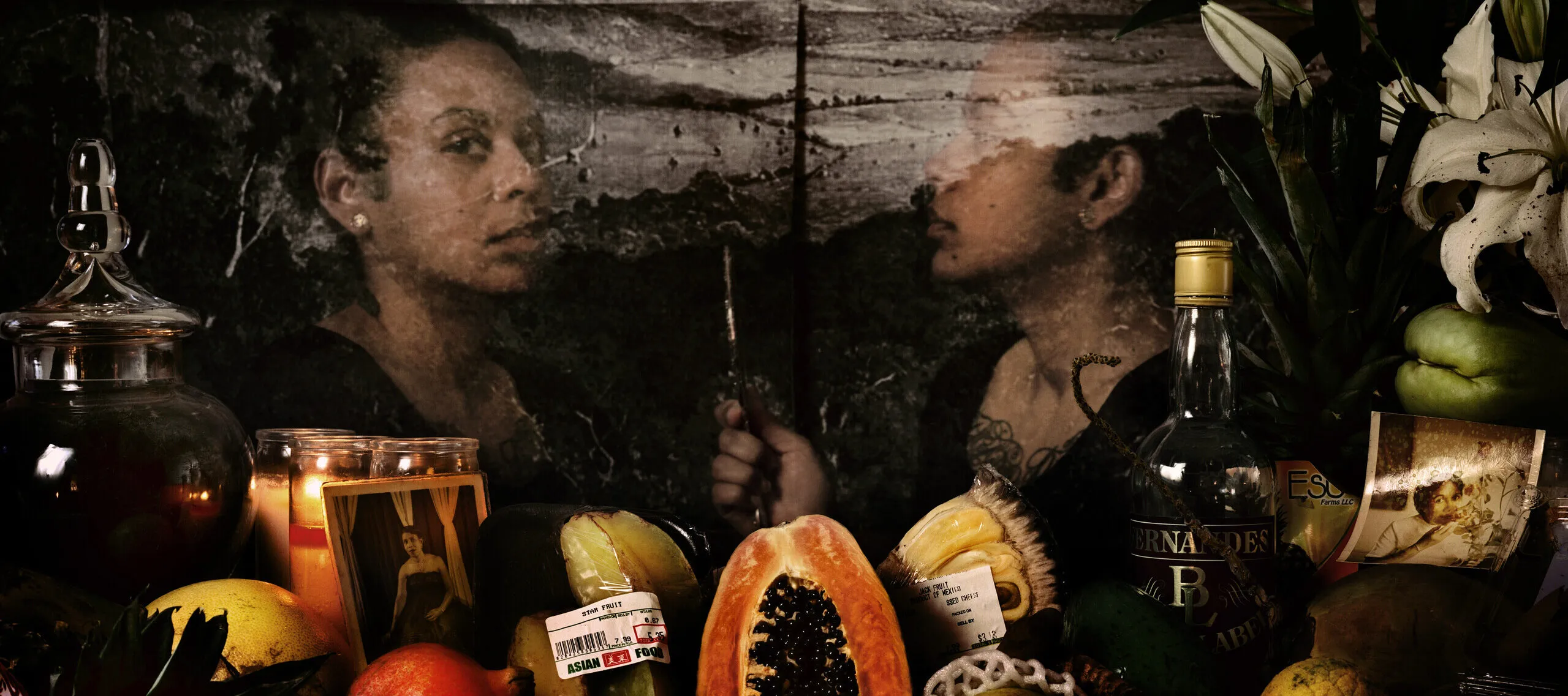 An assortment of flowers, colorful fruits, glass bottles, and photographs are visible in front of a transparent image of a dark-skinned woman who looks out at the viewer while her reflection looks at her. Behind her is a black-and-white image of a mountainous landscape.