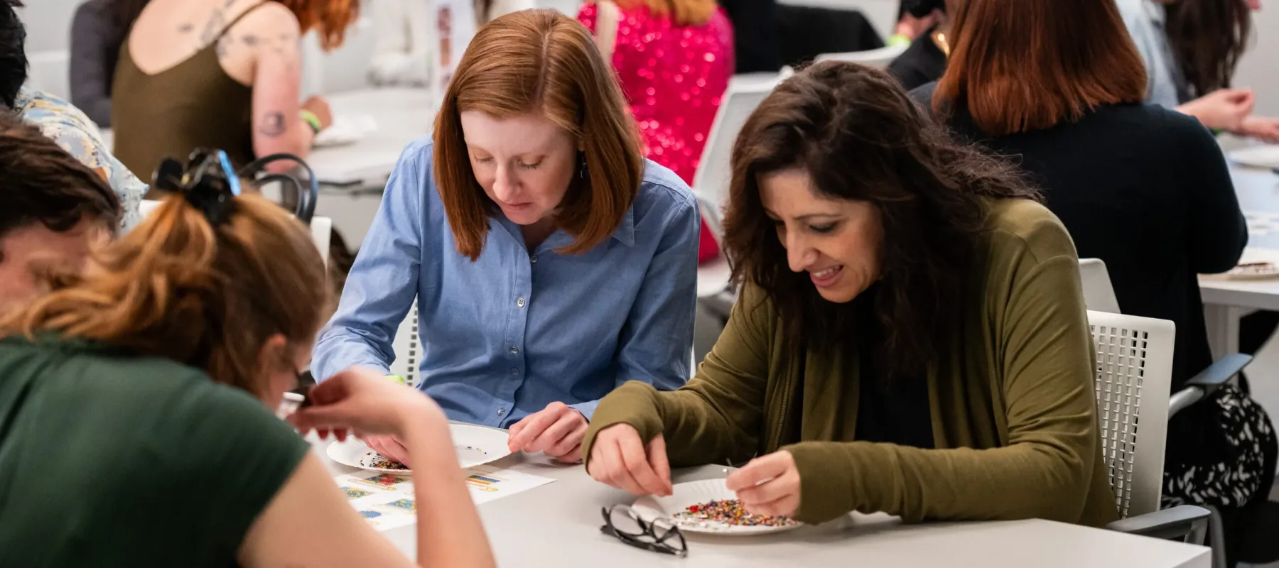 Numerous people sit at white tables, sorting through white, paper plates with multicolored breads and safety pins.