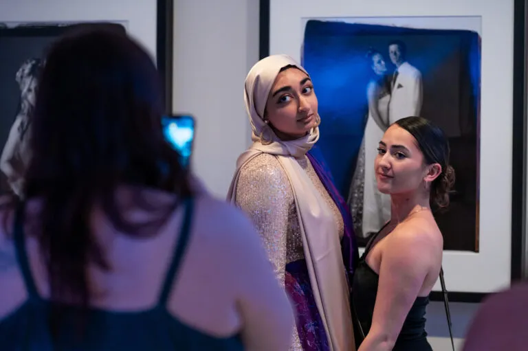 A girl with medium colored skin, a beige hijab, and a white and silver sequin outfit stands next to a girl with light-medium colored skin in a black dress. They are getting their photo taken with an iphone and stand in front of a black and white portrait photograph of a girl and boy in formal, prom wear.