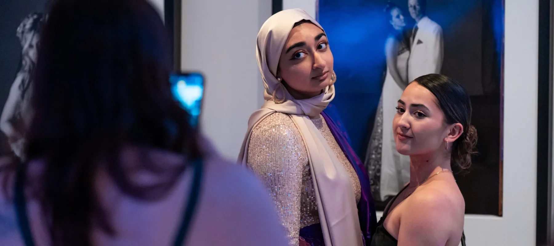 A girl with medium colored skin, a beige hijab, and a white and silver sequin outfit stands next to a girl with light-medium colored skin in a black dress. They are getting their photo taken with an iphone and stand in front of a black and white portrait photograph of a girl and boy in formal, prom wear.