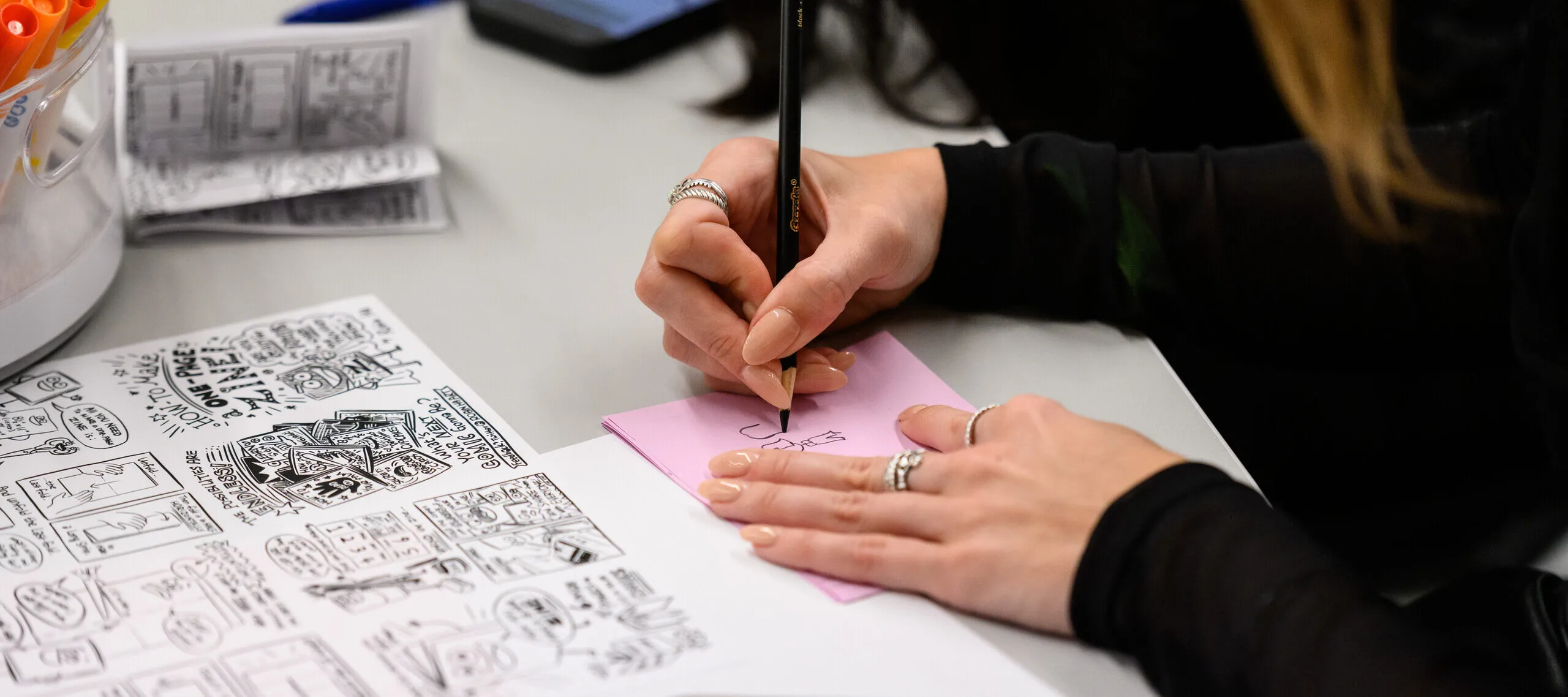 A person draws on a pink sheet of paper with another piece of paper with instructions alongside.