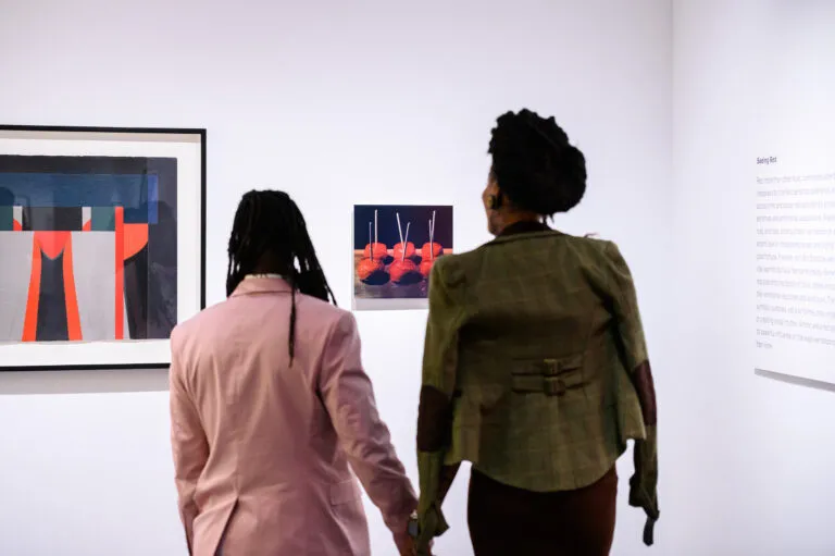 Two people, one in a pink jacket and one in an olive green jacket, hold hands and look at two artworks on a wall.