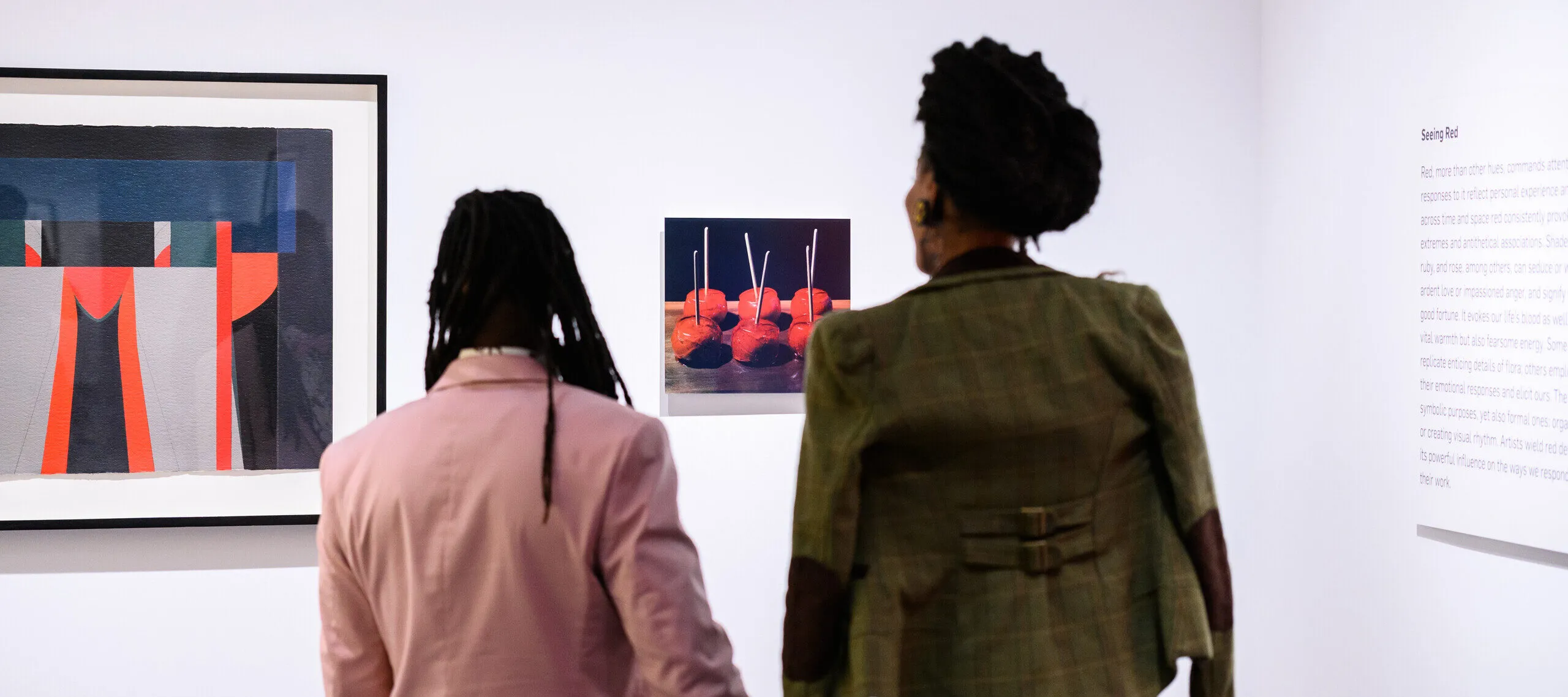 Two people, one in a pink jacket and one in an olive green jacket, hold hands and look at two artworks on a wall.