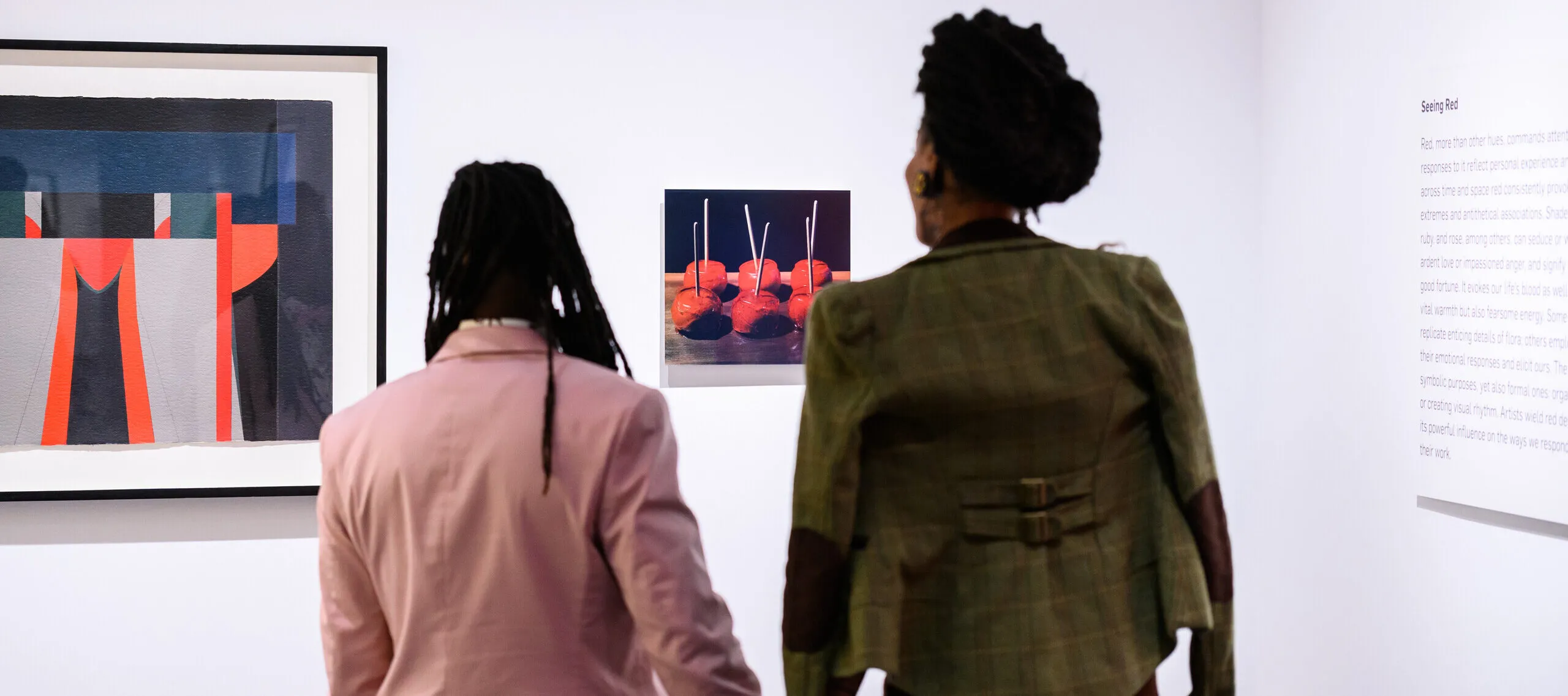 Two people, one in a pink jacket and one in an olive green jacket, hold hands and look at two artworks on a wall.