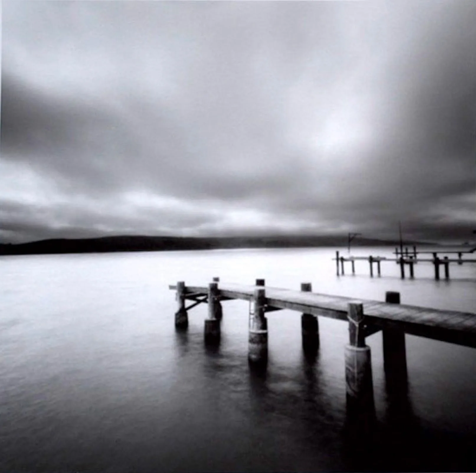 Soft focus black-and-white photograph of a still body of water with three parallel wooden docks to the right, receding into the background. Low hills hug the horizon line and an overcast sky caps off the image.