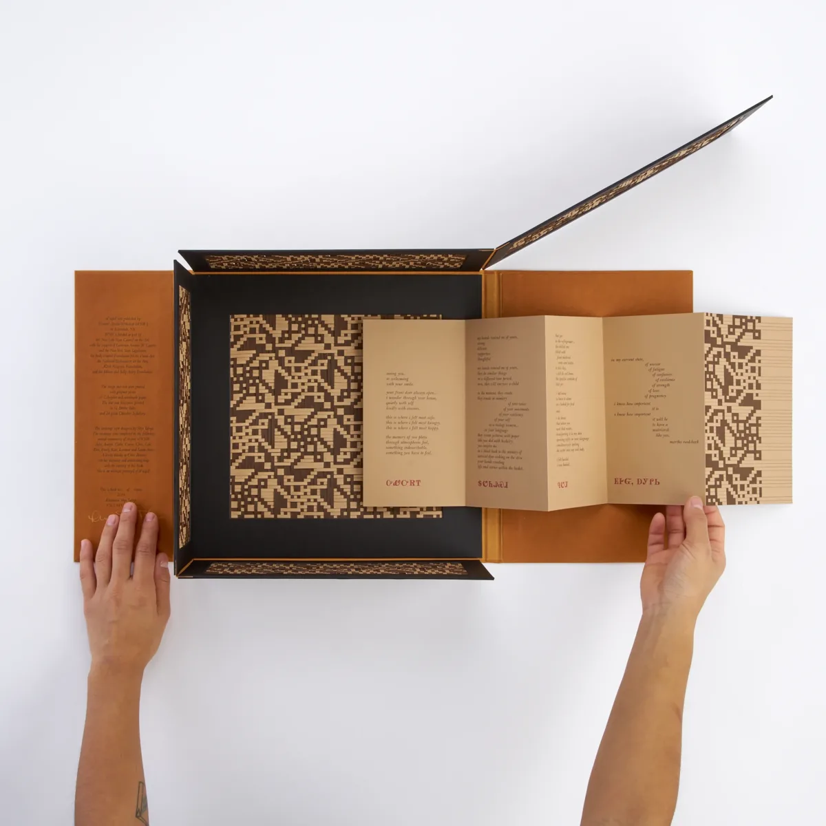 A geometric brown and beige patterned book with an unfolding cover reveals pages with printed text and patterns. The design includes symmetrical, abstract shapes and text in fold-out sections, laid out on a white surface.