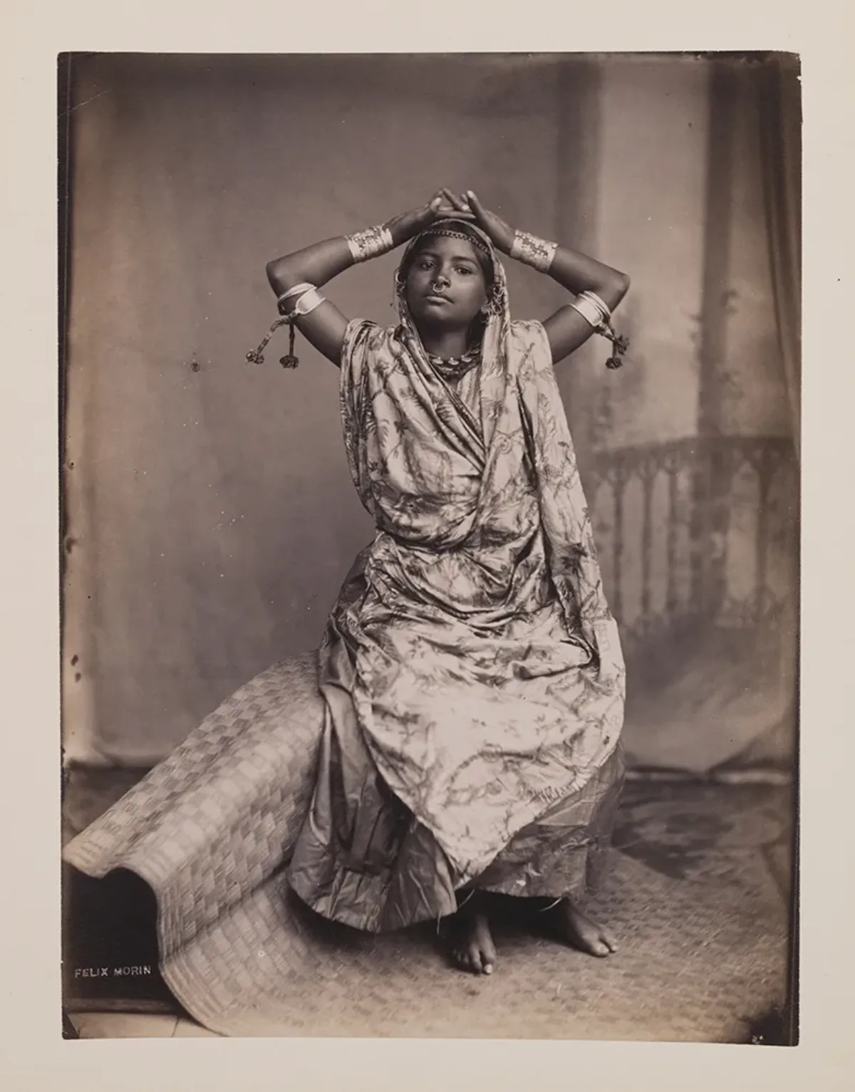 A black-and-white photograph shows a young dark-skinned woman in traditional Indian dress sitting on a woven mat. She places both hands atop her head, facing up. Her elbows are out to the side creating a diamond-like shape. She wears a nose ring, arm bangles, and a head adornment.