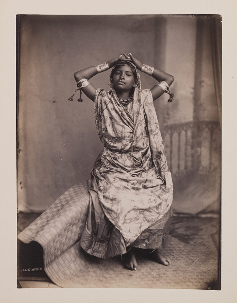 A black-and-white photograph shows a young dark-skinned woman in traditional Indian dress sitting on a woven mat. She places both hands atop her head, facing up. Her elbows are out to the side creating a diamond-like shape. She wears a nose ring, arm bangles, and a head adornment.