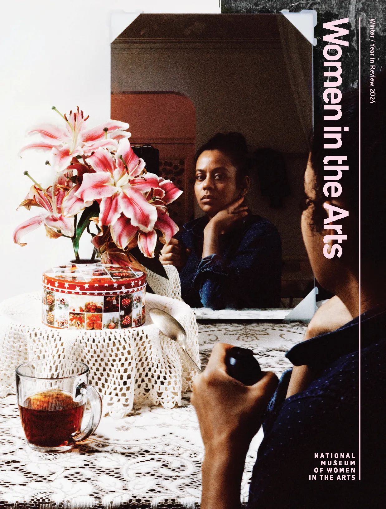 The cover of Women in the Arts Winter/Year in Review issue with an image of a dark-skinned woman looking at her reflection in the mirror. In front of her are pink flowers, a red and white round tin box, and a clear mug with dark brown liquid.