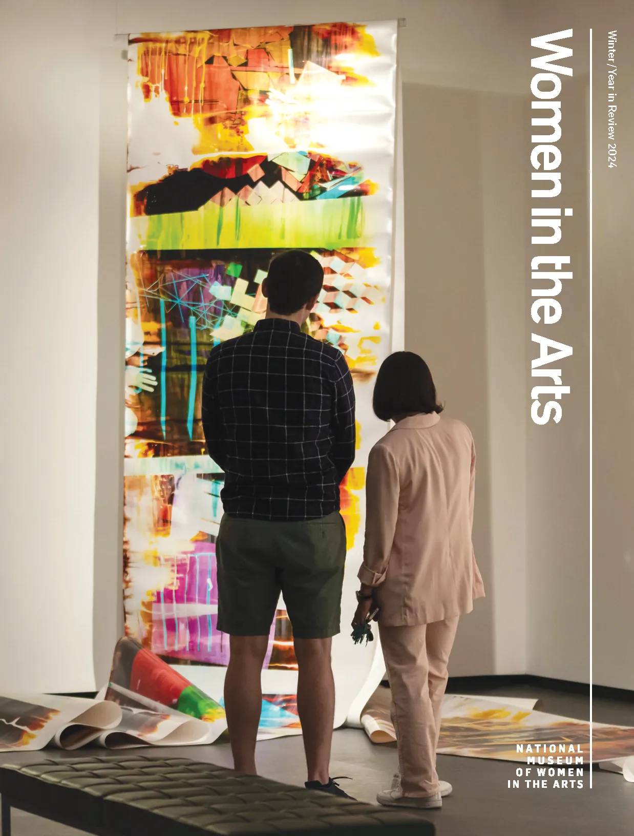 The cover of Women in the Arts year in review 2024 issue with two figures standing in front of a colorful abstract painting that is draped over a rod and falls in folds on the floor.