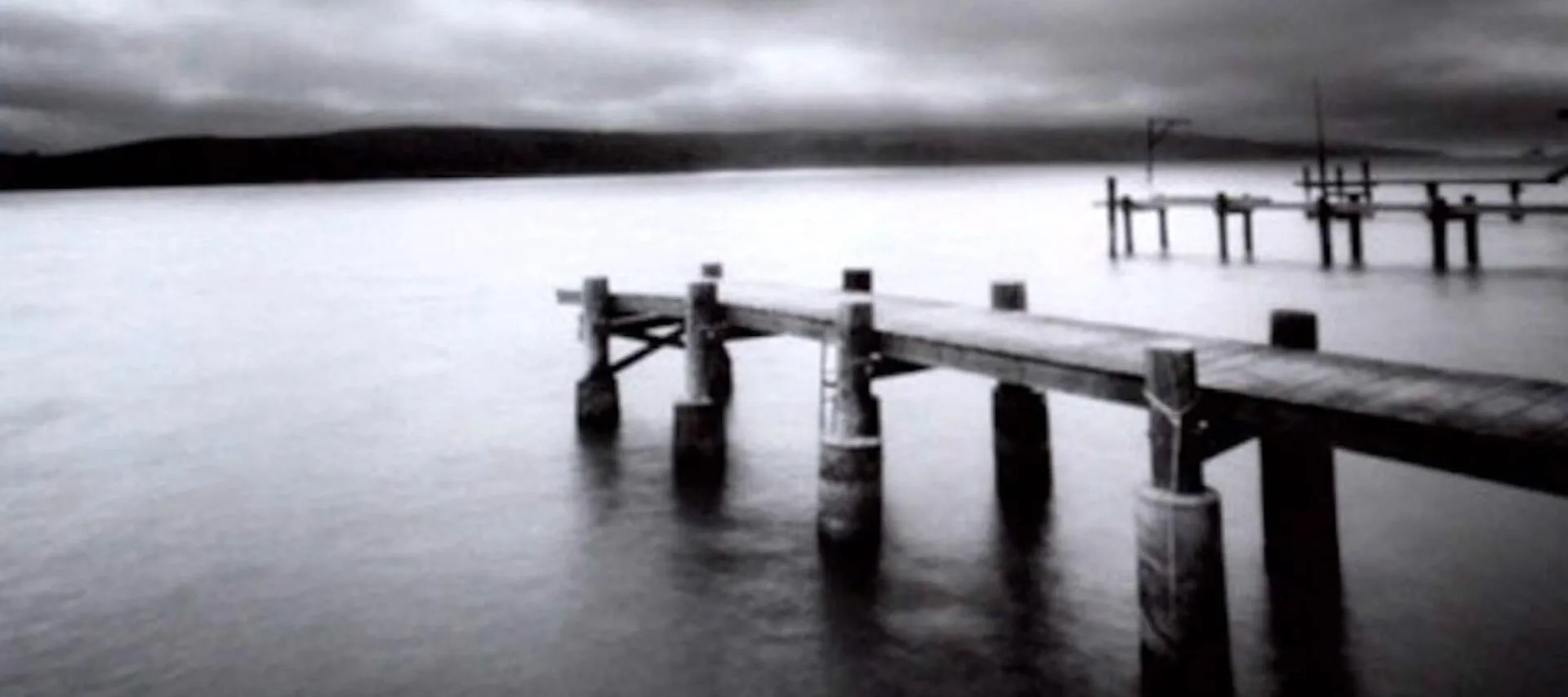 Soft focus black-and-white photograph of a still body of water with three parallel wooden docks to the right, receding into the background. Low hills hug the horizon line and an overcast sky caps off the image.