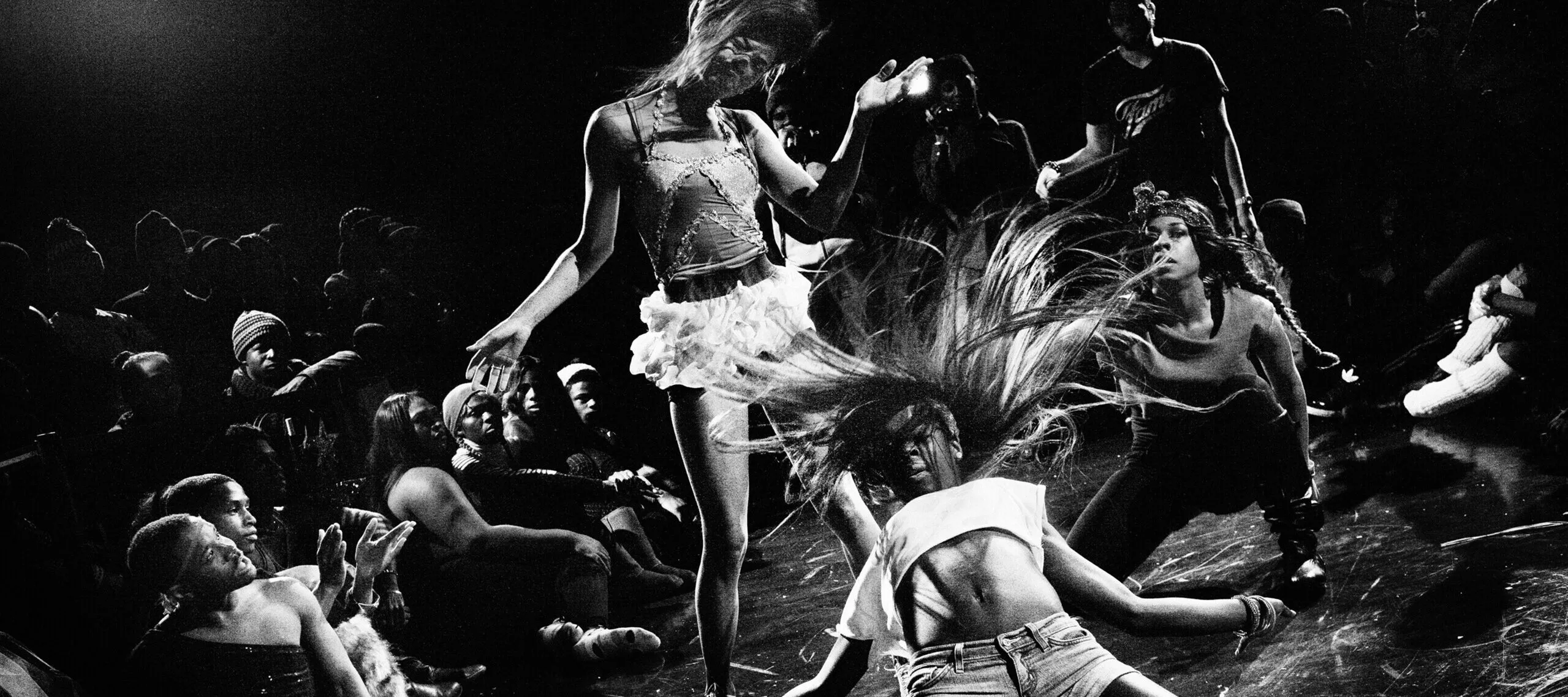 A black-and-white image shows a group of figures during a performance. The person in the foreground wears shorts and cropped shirt; they kneel on the ground with flailing hair while another person in a white skirt bends her head to one side. Other people stand around them and watch.