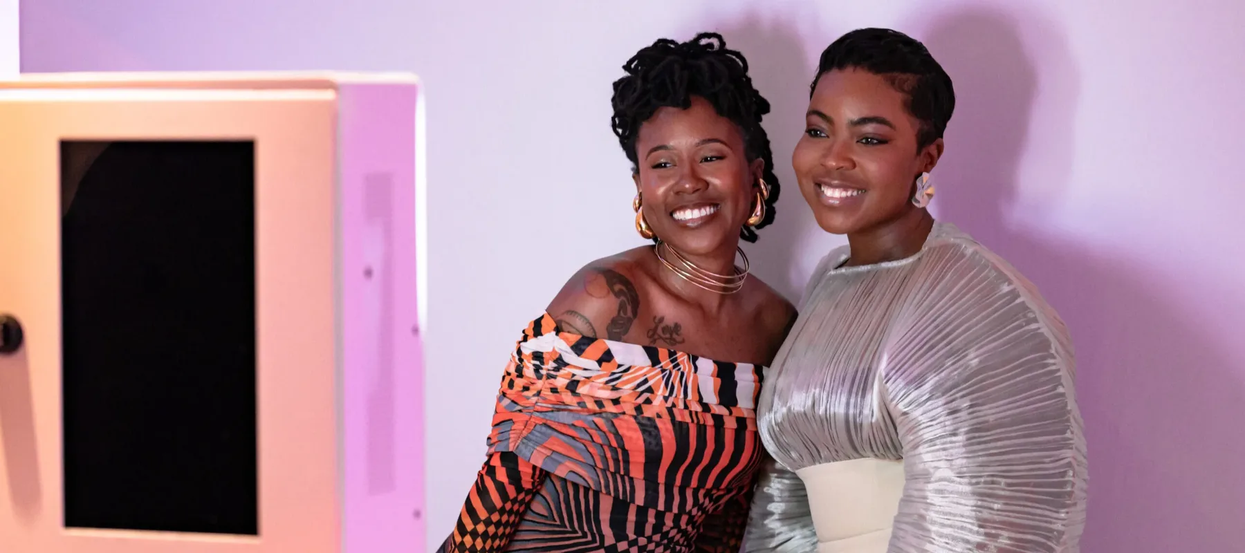 Two women with dark colored skin stand next to each other and smile in front of a white background for a photo. The woman on the left wears a gradient orange and purple dress with black stripes, and the woman on the right wears a silver and white dress.