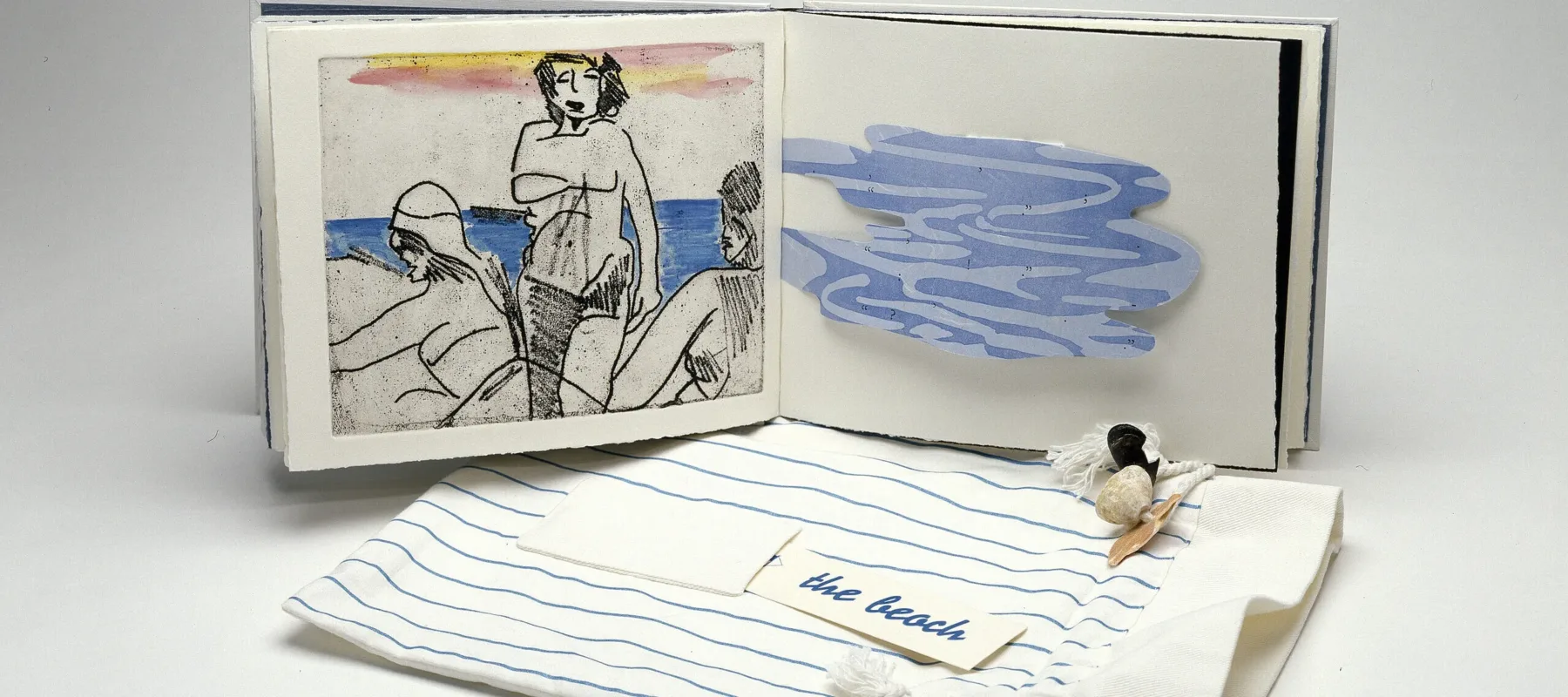 An open book is in the background and its cover is in the foreground. The left book page features a print of three figures on a beach and the right book page has a blue, amorphous shape with wavy lines. The book cover is a drawstring bag made of striped blue and white fabric. On top, it has a pocket that holds a card which reads “the beach” and is ornamented with seashells.