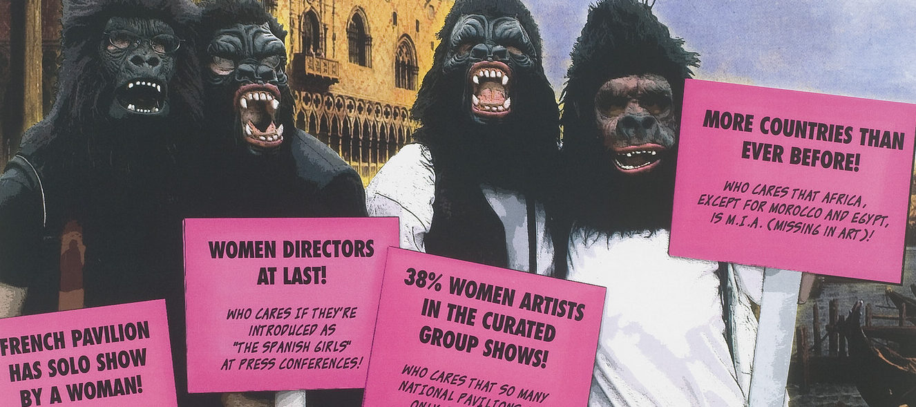 A poster shows four figures who are wearing gorilla masks and standing in front of a Venetian building. They hold pink picket signs. Red text beneath them reads, “Benvenuti alla Biennale Femminista!”