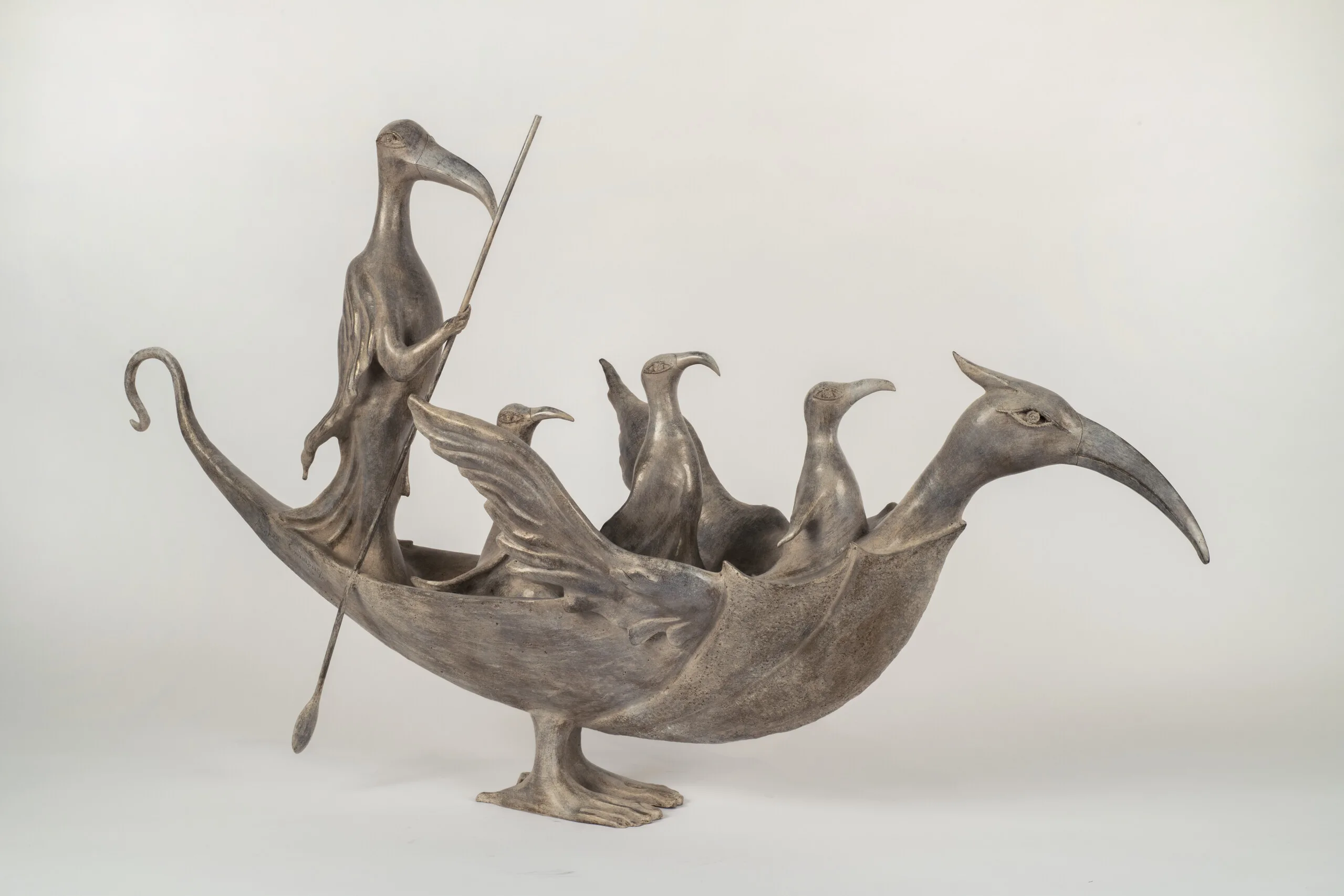 A bronze sculpture, seen from the side, merges the forms of a bird and a boat. The bird’s back is concave, forming the base of the boat, and its neck and wings extend outward. Several other, smaller bird-human hybrid creatures stand or sit within the boat like passengers in a ship. The tallest stands at the back, near the bird’s tail, cloaked in a robe and holding a long oar as if to steer the boat.