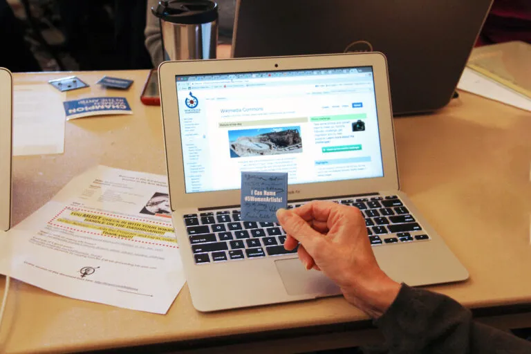 A hand holds a "I can name #5WomentArtists" pin in front of a laptop displaying the Wikimedia Commons website.