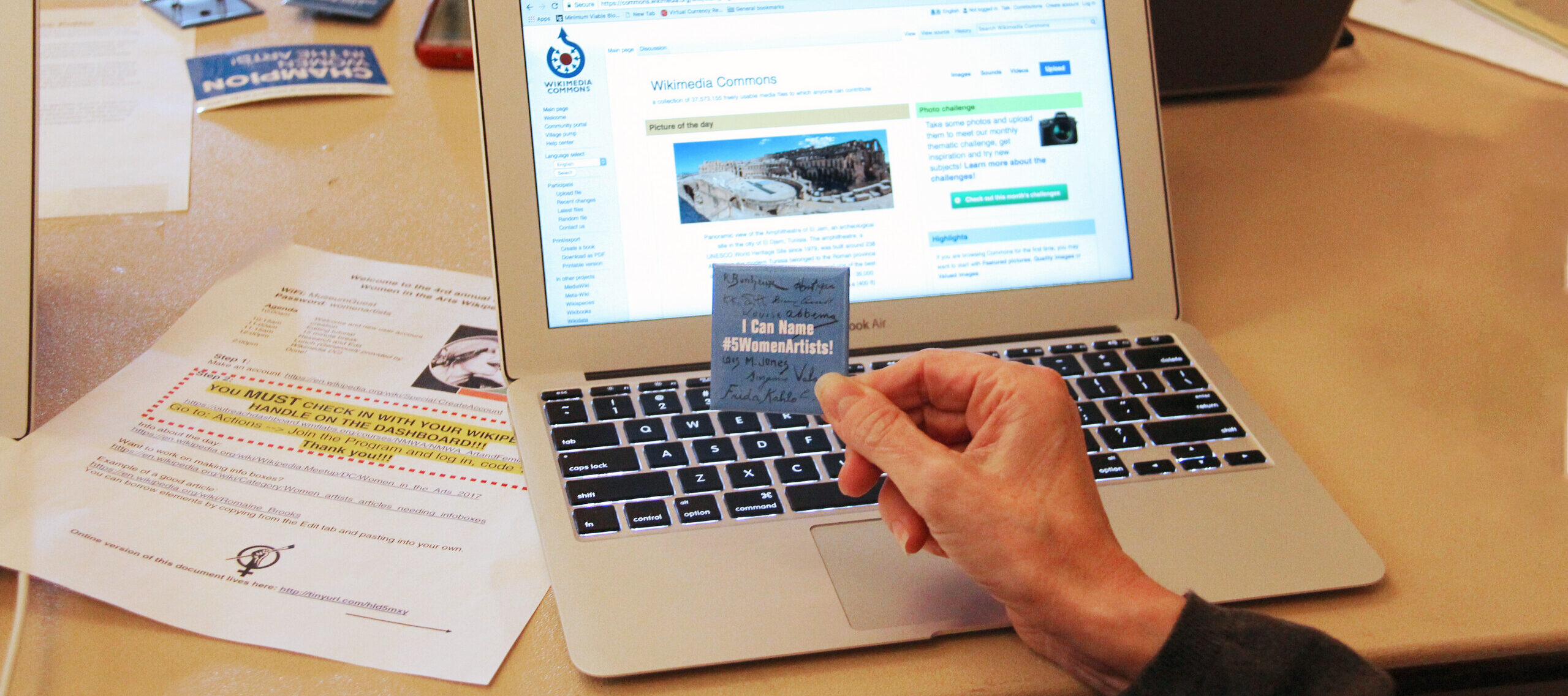 A hand holds a "I can name #5WomentArtists" pin in front of a laptop displaying the Wikimedia Commons website.