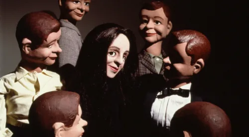 A light-skinned female ventriloquist dummy with straight black hair smiles in the center of a circle of six male dummy dolls with brown and dark hair.