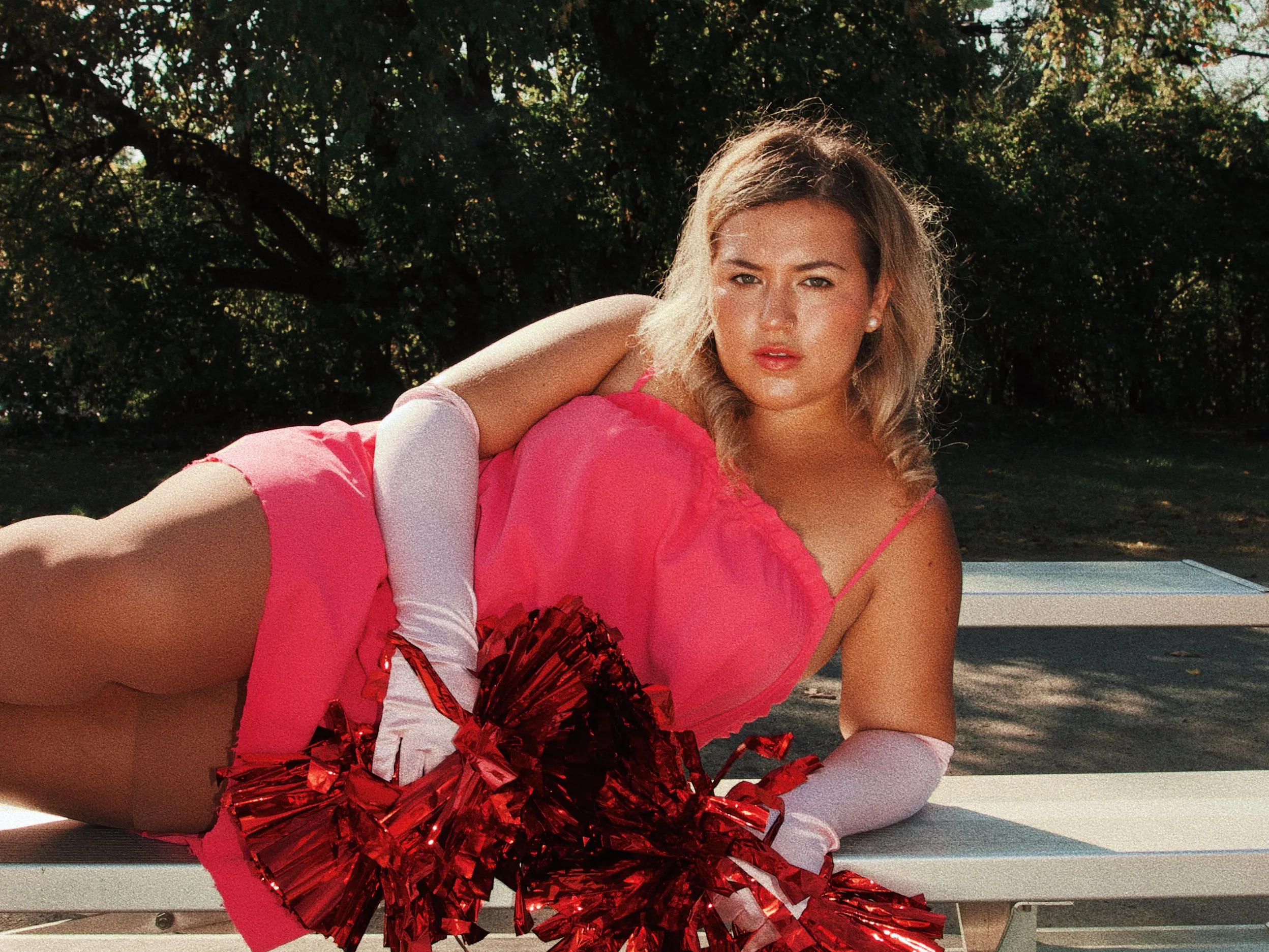 A woman smiles outdoors. She wears a coral dress, white gloves, and holds red pom-poms. The weather is sunny with a clear blue sky; trees and grass are in the background.