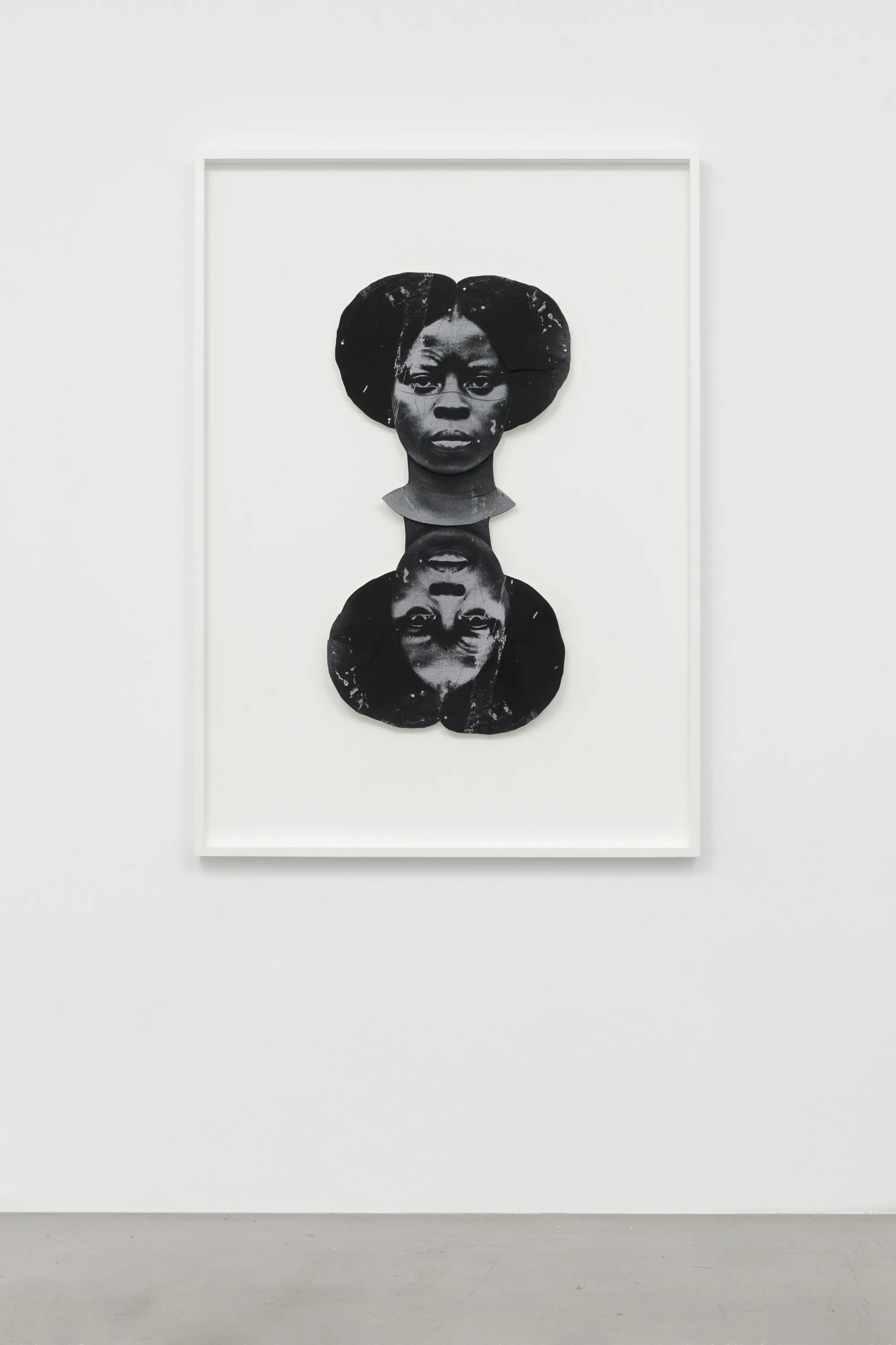 A black-and-white artwork features a dark-skinned woman's duplicated head, one facing the viewer straight on and the other upside down. The faces are stylized and abstract, with a textured appearance, set against a plain white background.