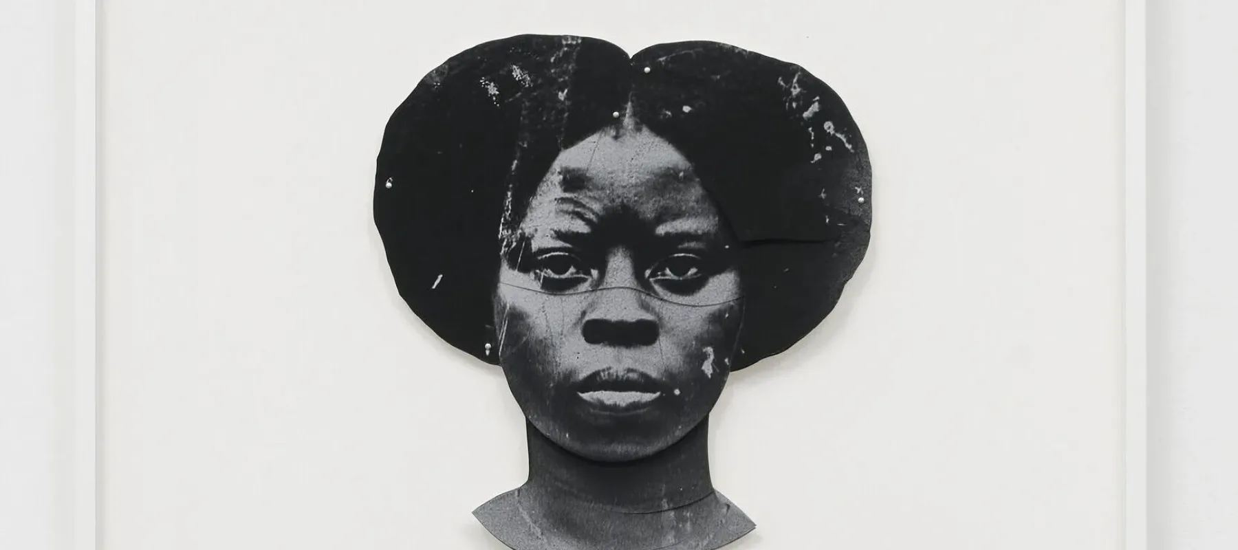 A black-and-white artwork features a dark-skinned woman's duplicated head, one facing the viewer straight on and the other upside down. The faces are stylized and abstract, with a textured appearance, set against a plain white background.