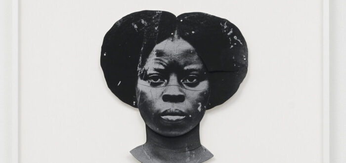 A black-and-white artwork features a dark-skinned woman's duplicated head, one facing the viewer straight on and the other upside down. The faces are stylized and abstract, with a textured appearance, set against a plain white background.