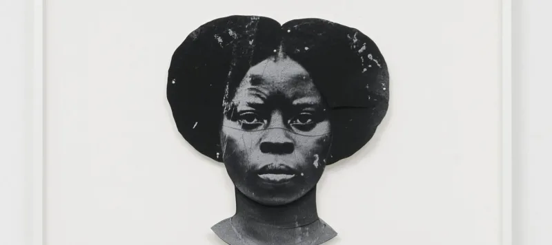 A black-and-white artwork features a dark-skinned woman's duplicated head, one facing the viewer straight on and the other upside down. The faces are stylized and abstract, with a textured appearance, set against a plain white background.