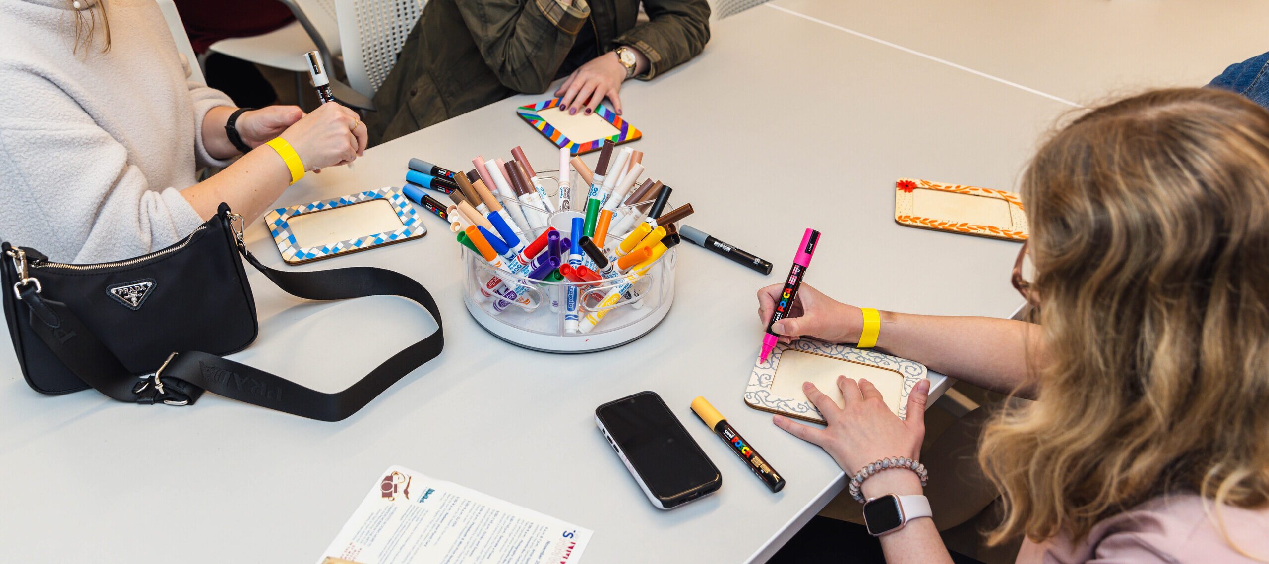 People are sitting around a table with art supplies, decorating picture frames. Several colorful markers are spread out, and a magazine is visible on the table. They appear engaged and focused on their creative activity.