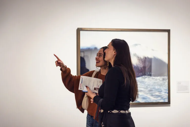 Two people stand in front of a framed artwork in a gallery. One person points towards the artwork while holding a brochure, and the other smiles, both engaged in conversation. The artwork appears to be a photograph with a water theme.