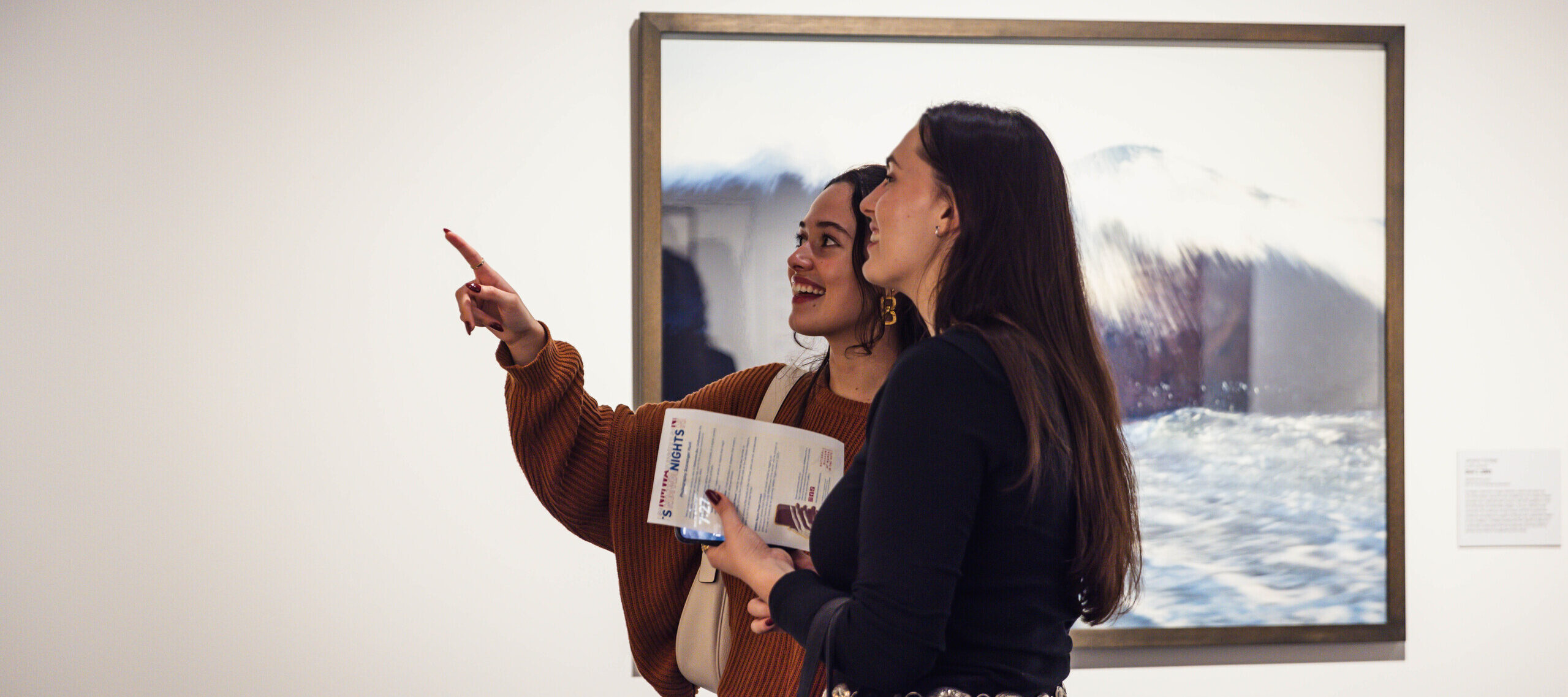 Two people stand in front of a framed artwork in a gallery. One person points towards the artwork while holding a brochure, and the other smiles, both engaged in conversation. The artwork appears to be a photograph with a water theme.