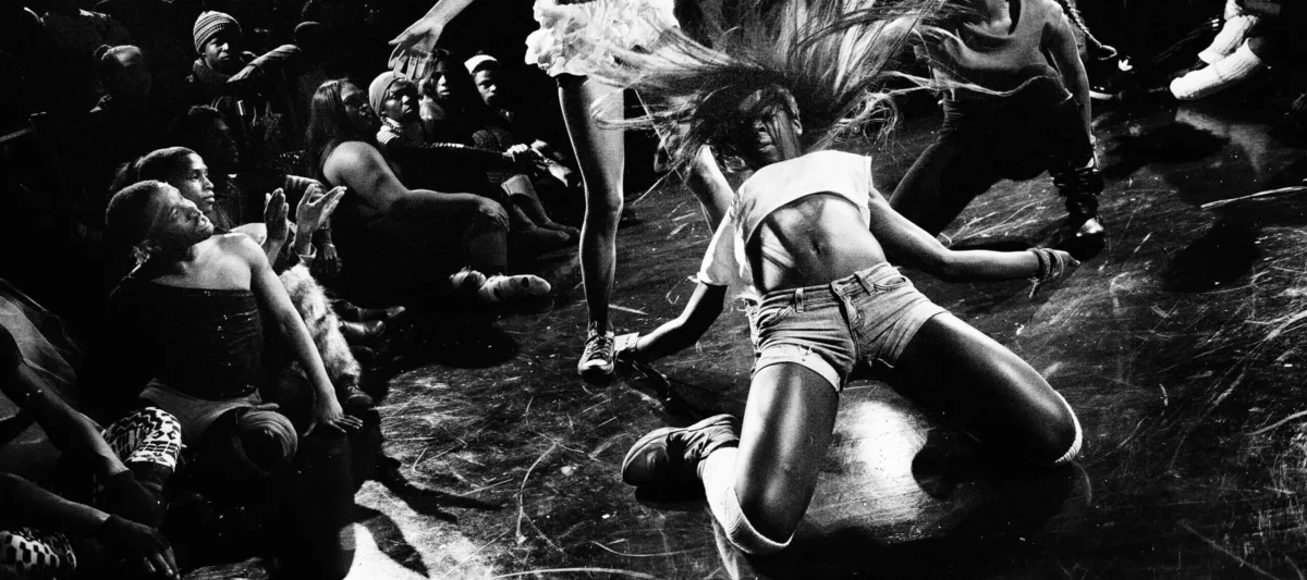 A black-and-white image shows a group of figures during a performance. The person in the foreground wears shorts and cropped shirt; they kneel on the ground with flailing hair while another person in a white skirt bends her head to one side. Other people stand around them and watch.