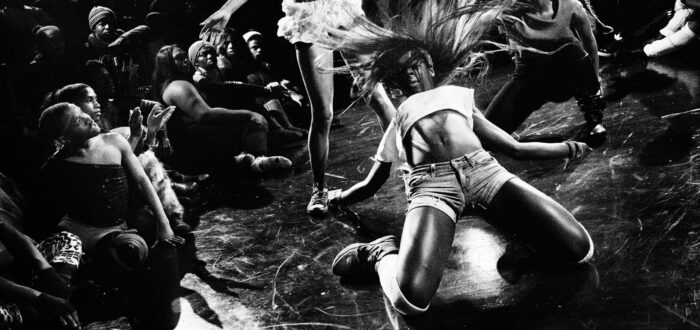 A black-and-white image shows a group of figures during a performance. The person in the foreground wears shorts and cropped shirt; they kneel on the ground with flailing hair while another person in a white skirt bends her head to one side. Other people stand around them and watch.