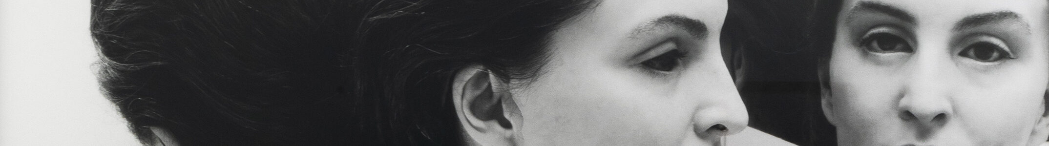 A light-skinned woman wears a realistic mask of another woman with the same skin tone. Her dark hair is pulled back into a low bun. She is dressed in a white tank top and surrounded by mirrors that reflect multiple angles of her face.