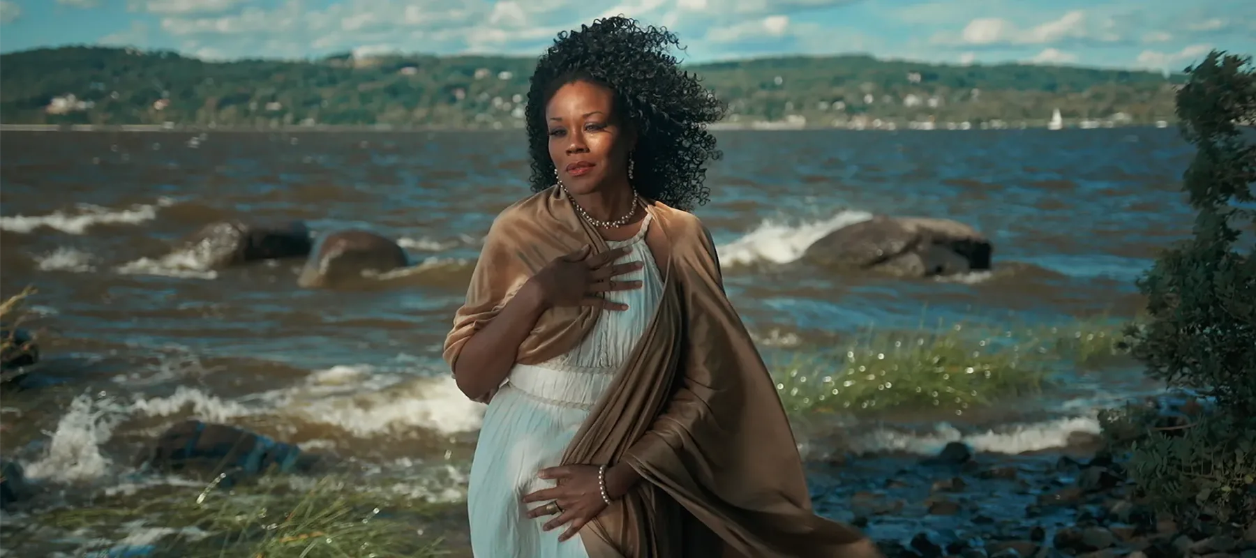 Standing gracefully on the sandy banks of the shore, a Black woman in a Grecian white robe embodies the mythic Aphrodite, posing as portrayed in Botticelli’s “Birth of Venus.”