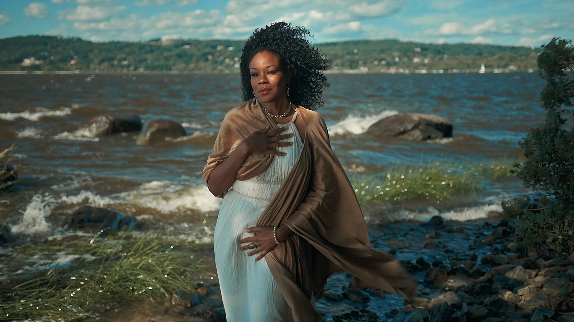 Standing gracefully on the sandy banks of the shore, a Black woman in a Grecian white robe embodies the mythic Aphrodite, posing as portrayed in Botticelli’s “Birth of Venus.”