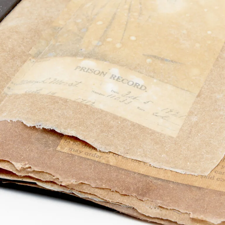 A  detailed view of a box-shaped book featuring aged, hand cut paper, layered with torn edges. The words "prison record" can be seen through the semi-translucent pages.