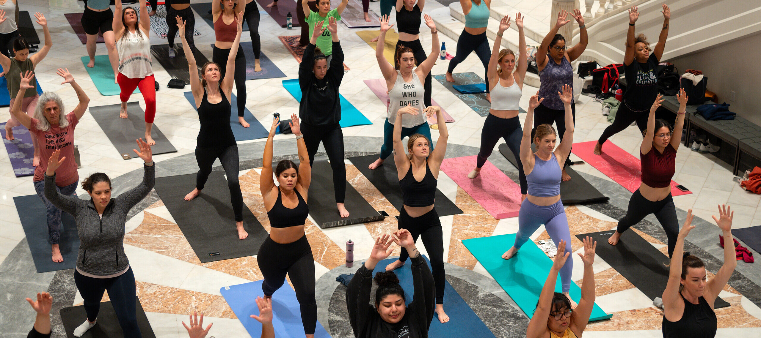 A crowd participates in a yoga class in the Great Hall of the National Museum of Women in the Arts. They all wear athletic apparel and are standing on their own yoga mats in a "warrior 2" pose.