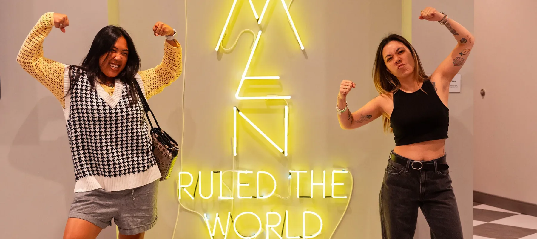 Two woman flex their arm muscles in front of a yellow neon sign that reads "what if women ruled the world"