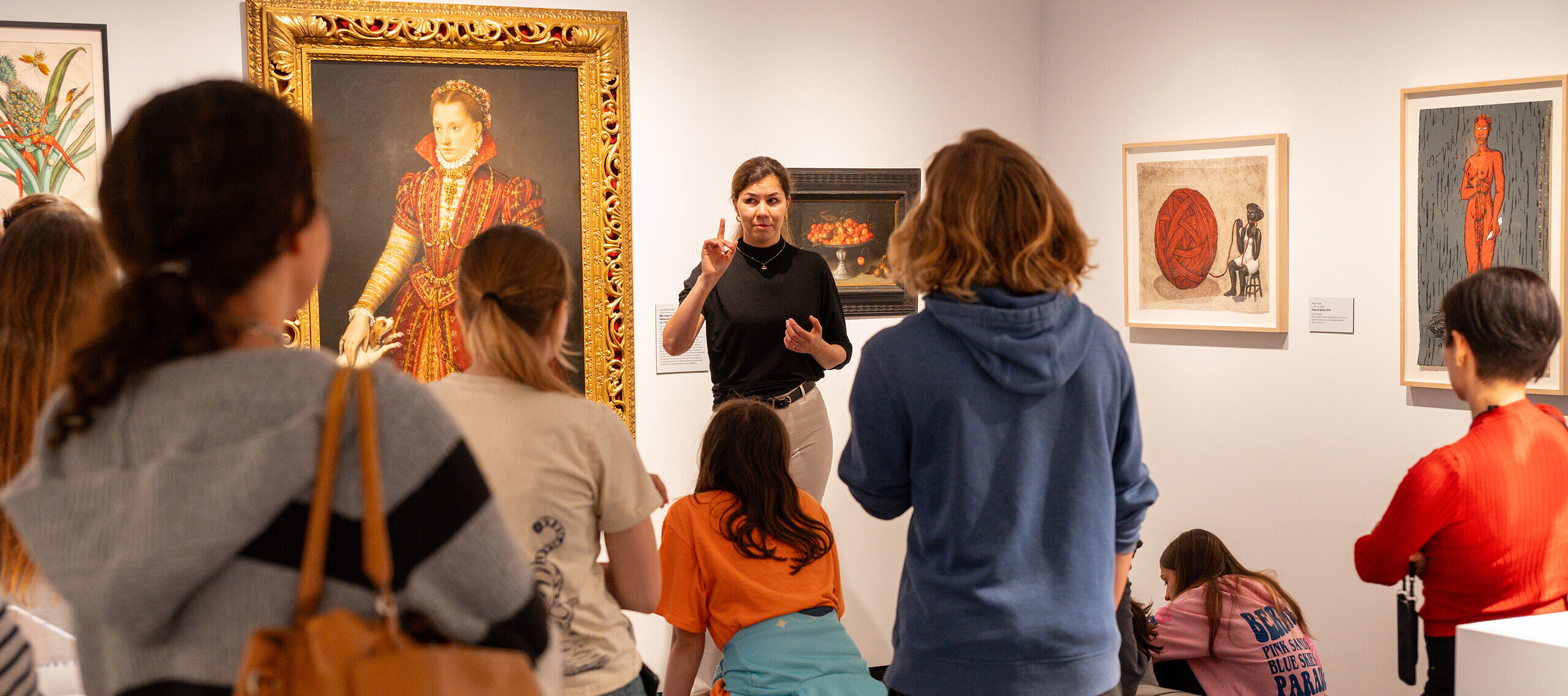 A woman with light colored skin, wearing a black top and white pants, talks to a group of people in a gallery room. She stands in front of a large, painted portrait of a woman in a high-collared red dress.