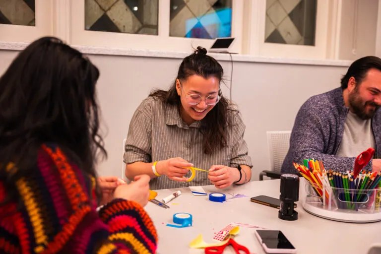 Three people sit at a grey table, decorating paper bookmarks. In the middle, a woman with light-medium skin, brown hair, and clear glasses laughs as she pulls yellow tape over her bookmark.