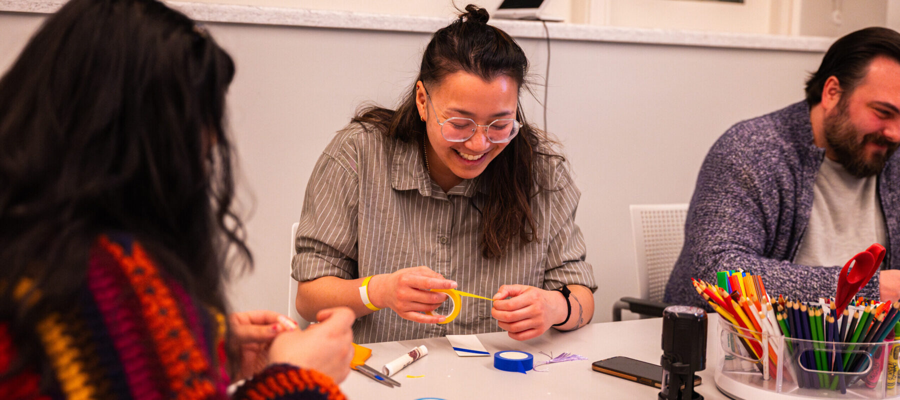 Three people sit at a grey table, decorating paper bookmarks. In the middle, a woman with light-medium skin, brown hair, and clear glasses laughs as she pulls yellow tape over her bookmark.