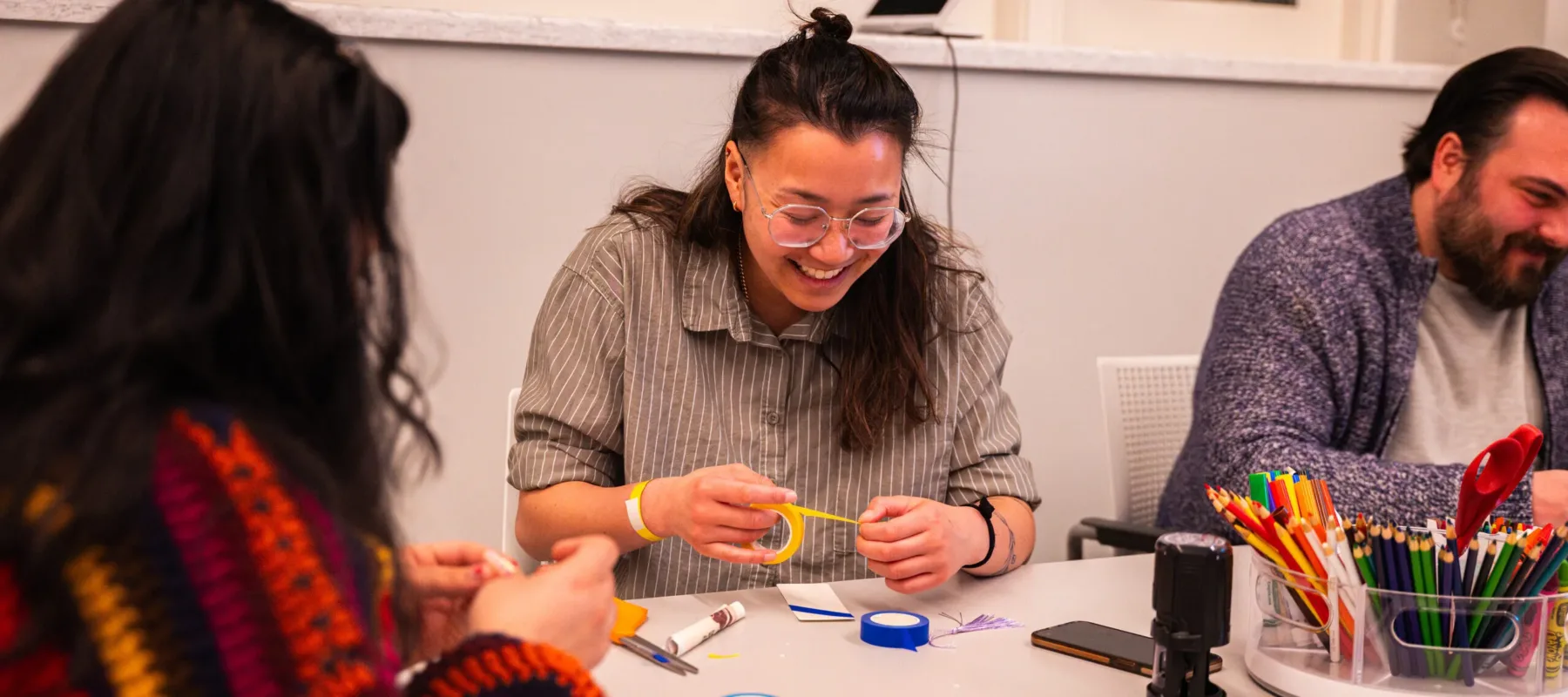 Three people sit at a grey table, decorating paper bookmarks. In the middle, a woman with light-medium skin, brown hair, and clear glasses laughs as she pulls yellow tape over her bookmark.