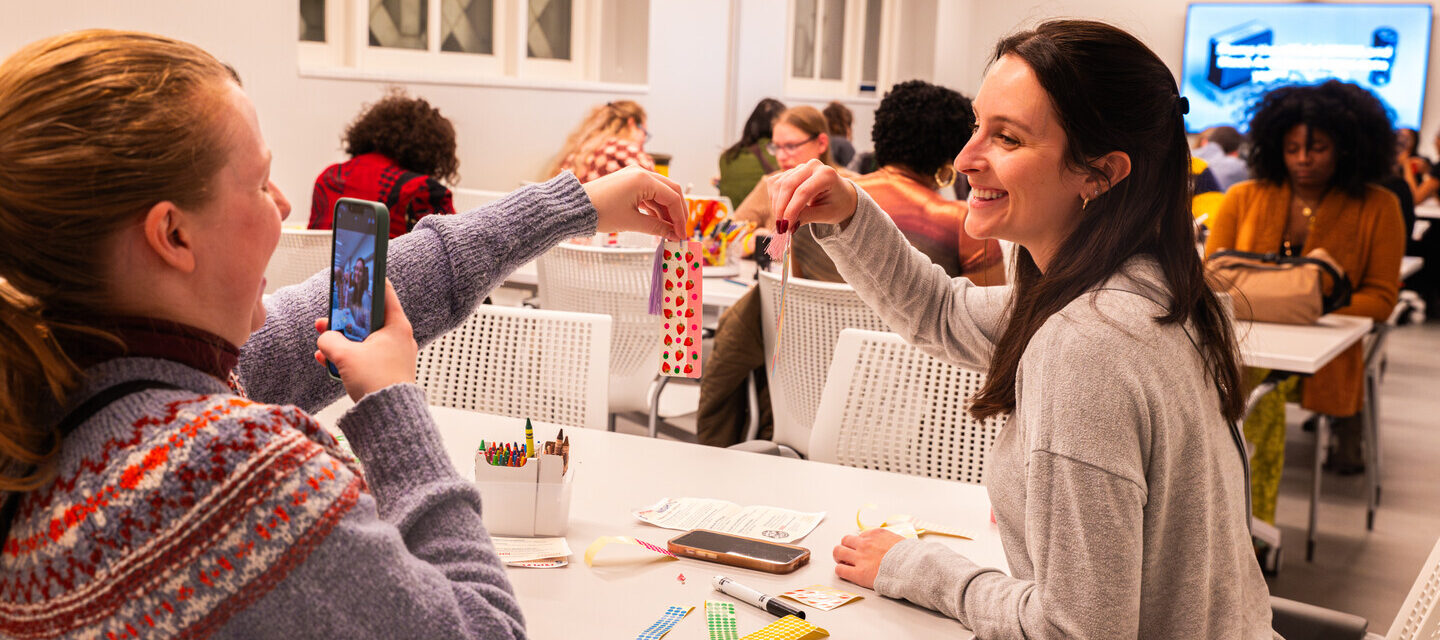 Two people with light skin tone hold up decorated bookmarks next to each other. The person on the left takes a picture of the bookmarks with her iPhone.