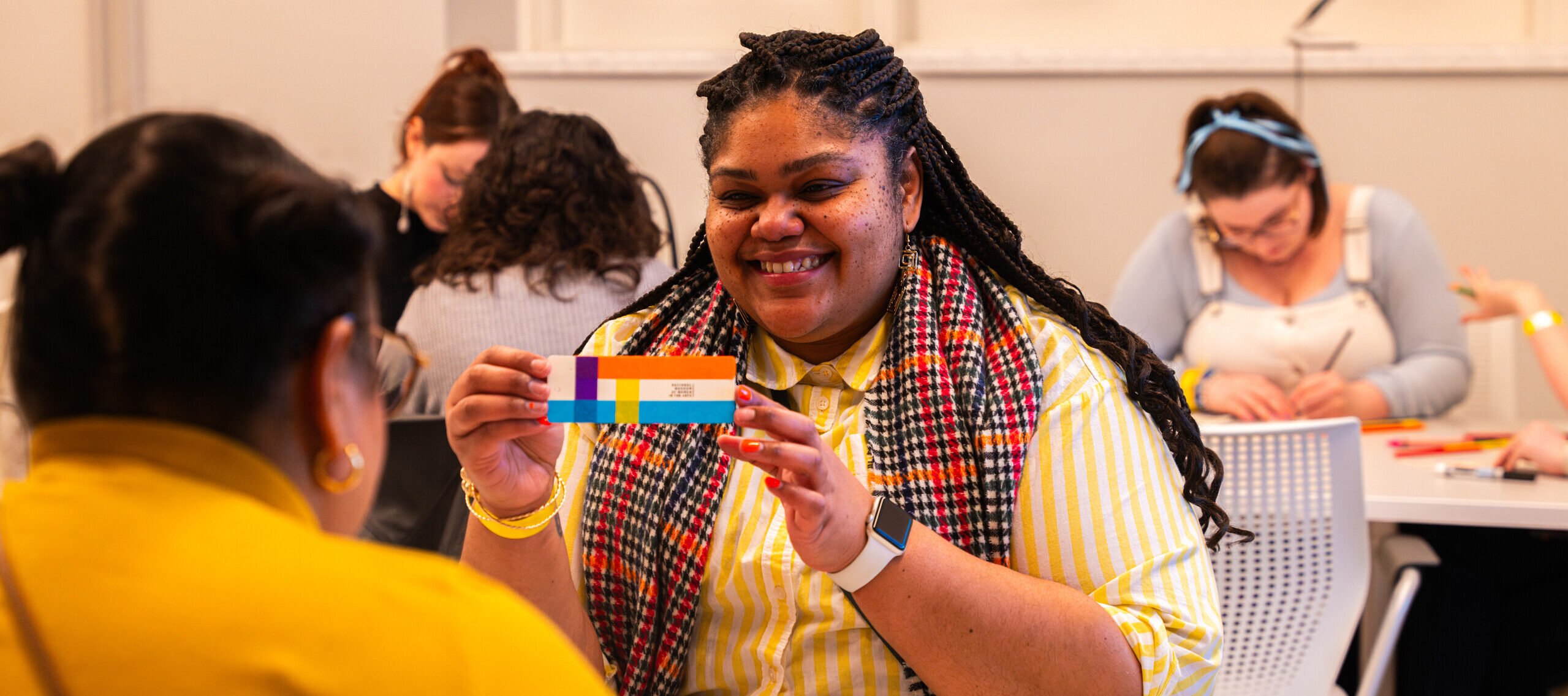 A woman with dark colored skin sits at a grey table. She wears a white and yellow striped sweater and a black, red, and orange plaid scarf. She holds a striped bookmark and smiles at a person across the table from her.