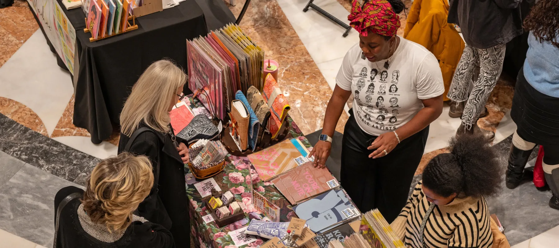 An aerial shot of two people looking at brightly colored notebooks, stickers, and prints. Two people behind the table selling the items. 