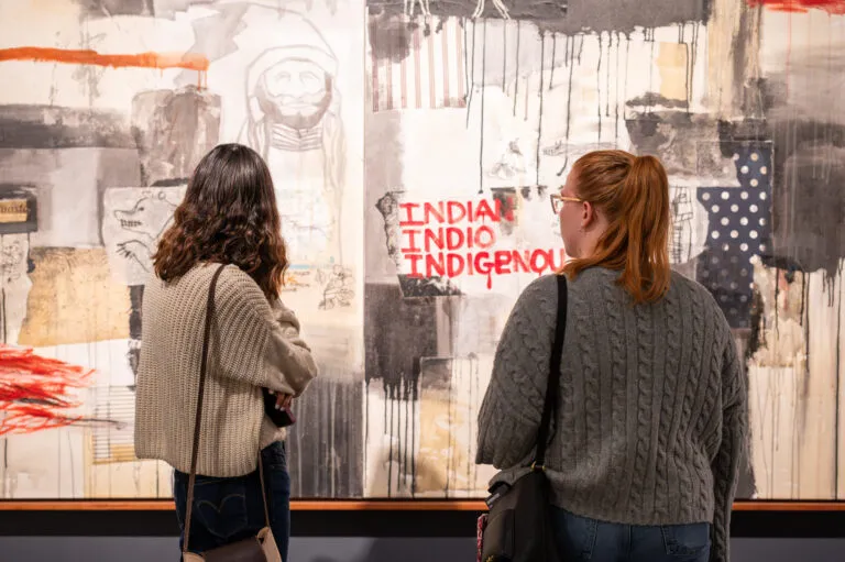 Two female visitors in grey sweaters look at an abstract painting. The painting is primarily beige and grey blocks of color, and the words "Indian" "Indio" and "Indigenous" are written in red paint.
